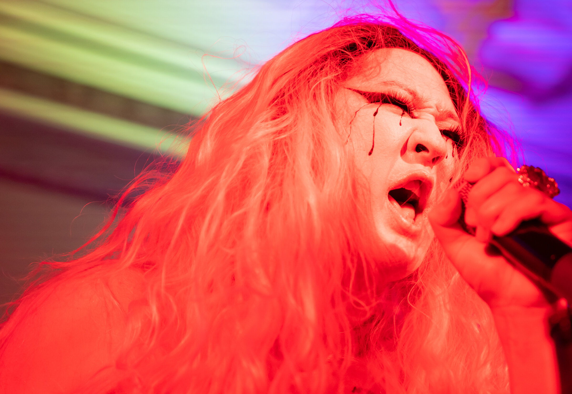 Close-up of a person singing into a microphone, bathed in red light. Black streaks run from their closed eyes. Background is a blur of green and purple.